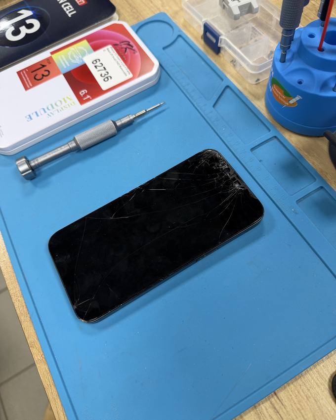 iPhone 13 with a cracked screen on the repair mat, next to a screwdriver and the replacement display box